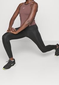 Personne en legging noir et débardeur marron effectuant un étirement en fente avant avec les mains jointes au-dessus du genou fléchi.