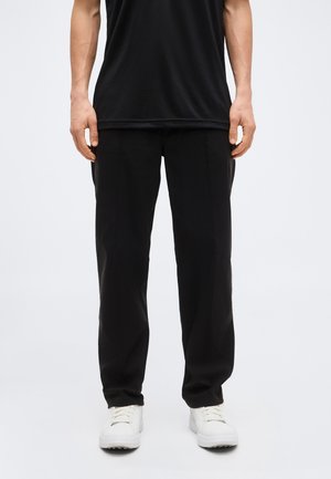 Άτομο που φοράει μαύρο παντελόνι, μαύρο πουκάμισο και λευκά αθλητικά παπούτσια, στέκεται μπροστά σε έναν απλό ανοιχτόχρωμο φόντο.