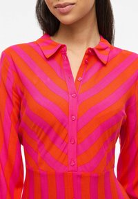 Chemise boutonnée avec des rayures verticales en rose vif et orange. Elle possède un col pointu et des boutons le long de la patte de boutonnage avant.