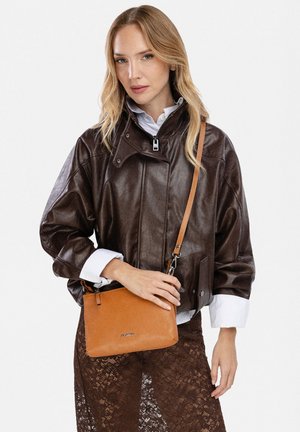 Femme portant une veste en cuir marron foncé, une chemise blanche et une jupe marron à motif dentelle, tenant un sac bandoulière en cuir beige.