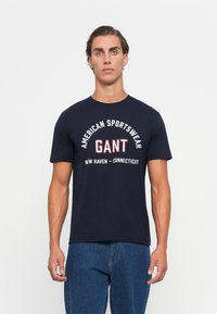 Bărbat stând drept, îmbrăcat cu un tricou GANT bleu marin pe care scrie "American Sportswear" și blugi, pe un fundal simplu deschis.