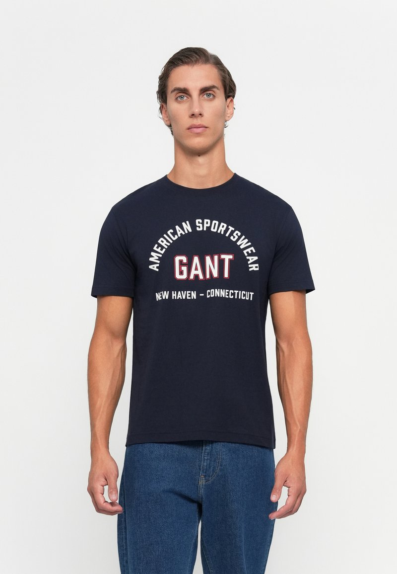 Vyras stovi tiesiai, vilkėdamas tamsiai mėlyną GANT marškinėlį su užrašu "American Sportswear" ir mėlynas džinsus, priešoveikį šviesiai neutralų foną.