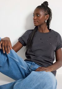 Grå t-shirt med oprullede ærmer, prydet med spredte sorte udsmykninger. Båret med løse, lys blå denim jeans.