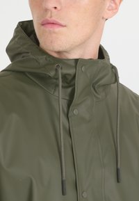 Olivgrüne, wasserdichte Jacke mit hohem Kragen, Frontdruckknöpfen und verstellbarem Kordelzug. Hat eine glatte, glänzende Textur und keine Muster.