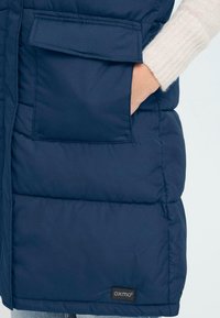 Tums zilas polsterēts veste ar izšūtu dizainu, kurai ir sānu kabata un zīmola logo etiķete. Audums izskatās gluds un izturīgs.