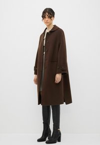 Cappotto in lana marrone con bottoni grandi e tasche laterali, abbinato a stivali in pelle nera al ginocchio e a una gonna con motivo sotto.
