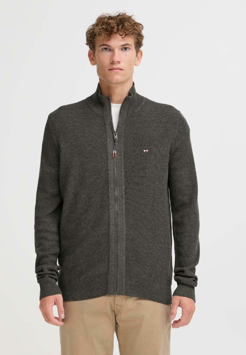 Mørkegrønn zip-up genser med teksturert strikk, høy krage og ribbestrikkede mansjetter. Har en liten logo-lapp på brystet.