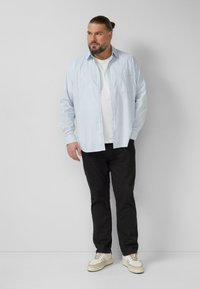 Hellblaues Hemd mit Knöpfen, weißes T-Shirt darunter, schwarze Hose und beige Turnschuhe. Verfügt über eine einzelne Brusttasche, legeren Schnitt und lange Ärmel.