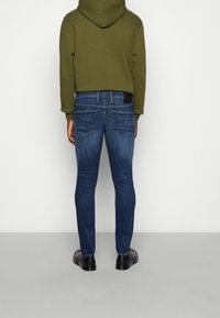 Slim-fit jeans i mörkblå denim med subtila blekningar. Bakifrån syns två bakfickor och en svart etikett vid midjebandet. Matchas med en grön hoodie.