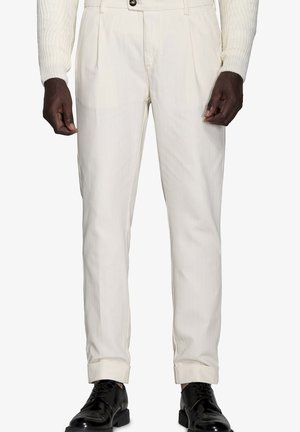 Helle, plissierte Hose mit geradem Schnitt, aus Baumwollstoff, und aufgerollten Säumen, kombiniert mit polierten schwarzen Schuhen.
