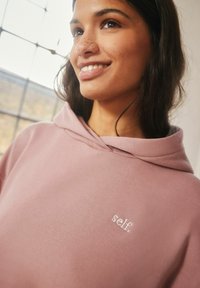 Roosa kapuutsiga sviiter, mis on valmistatud pehmest kangast, millel on kaeluse lähedal peenembroidery logo "self." Sujuv tekstuur ja mugav disain.
