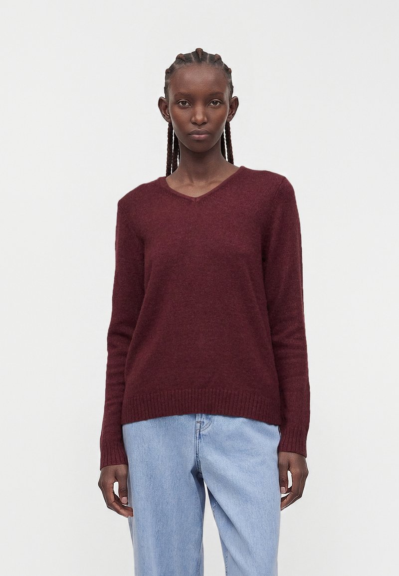 Femme aux cheveux tressés portant un pull bordeaux uni à col en V et un jean bleu clair, debout devant un fond blanc.