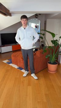 Let jakke med hvid lynlås over en sort skjorte, grå ternede bukser og hvide sneakers. Indendørs omgivelser med en grøn plante og en orange sofa.