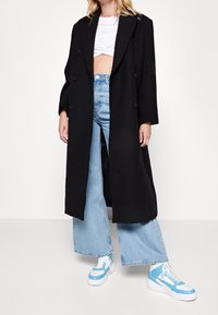 Abrigo largo de mezcla de lana negra con diseño de doble botonadura, combinado con unos vaqueros de pierna ancha en azul claro y zapatillas blancas con detalles en azul.