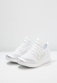 Vita atletiska skor med ett ventilerande mesh-övre, silverdetaljer på tån och en texturerad gummisula. Skosnören och logotyp är synliga.