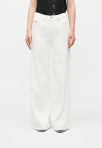 Άτομο φοράει λευκό ψηλόμεσο παντελόνι με φαρδιά μπατζάκια, συνδυασμένο με μαύρο αμάνικο πουκάμισο με κουμπιά, στέκεται με τα χέρια χαλαρά στο πλάι.