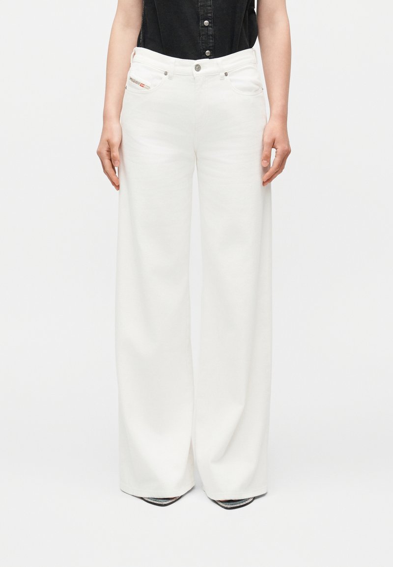 Άτομο φοράει λευκό ψηλόμεσο παντελόνι με φαρδιά μπατζάκια, συνδυασμένο με μαύρο αμάνικο πουκάμισο με κουμπιά, στέκεται με τα χέρια χαλαρά στο πλάι.