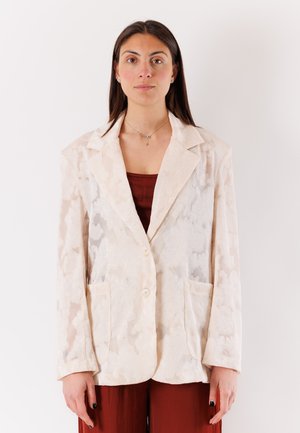 Donna con lunghi capelli scuri indossa un blazer crema a motivi sopra una maglia rosso scuro e pantaloni abbinati, in piedi davanti a uno sfondo bianco semplice.