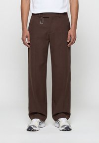 Άνδρας που φοράει χαλαρά καφέ παντελόνια με ένα καραμπίνερ στο ζωνάρι, λευκά αθλητικά παπούτσια και λευκό μπλουζάκι μέσα στην παντελόνα.