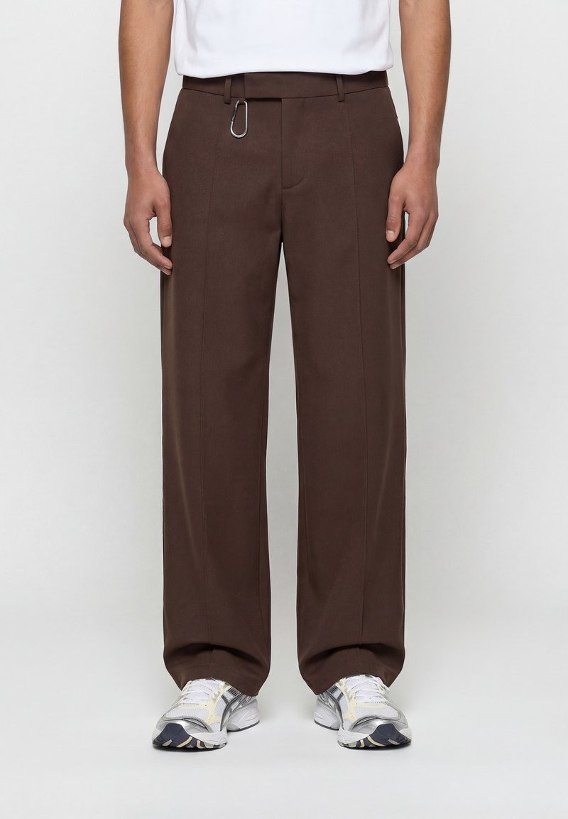 Άνδρας που φοράει χαλαρά καφέ παντελόνια με ένα καραμπίνερ στο ζωνάρι, λευκά αθλητικά παπούτσια και λευκό μπλουζάκι μέσα στην παντελόνα.