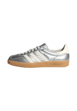 Baskets Adidas Gazelle argentées avec des bandes blanches, une languette de talon blanche, une semelle en gomme et une fermeture à lacets, montrées de profil sur fond blanc.