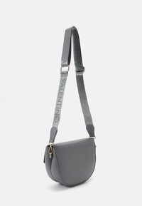 Šedá kožená crossbody taška s zakřiveným tvarem. Obsahuje popruh s vyraženým logem, zlaté kovové detaily a hladkou texturu.