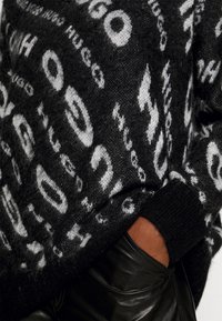 Zwarte gebreide trui met een pluizige textuur en witte merkpatronen. Zwarte geribbelde manchetten. Zwarte leren broek zonder kreukels zichtbaar.