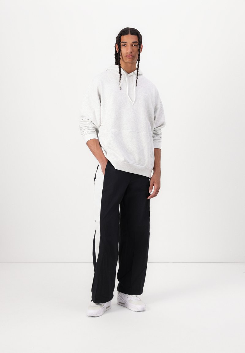 Grauer Kapuzenpullover mit Kängurutasche, kombiniert mit schwarzen Hosen mit weißen Seitenstreifen. Das Modell steht in weißen Sneakers vor einem schlichten Hintergrund.