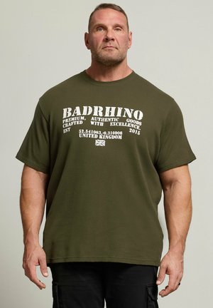 Muscular man wearing an olive green "BADRHINO" t-shirt with white text and coordinates, standing against a plain light background.