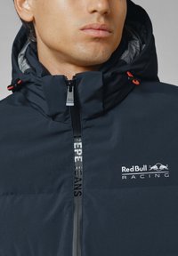 Tamnomo modra jakna na zadrgo s kapuco, ki ima rdeče detajle, logo "Red Bull Racing" in blagovno znamko "Pepe Jeans" na zadrgi. Gladka tekstura.