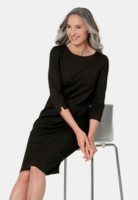 Robe noire avec des manches trois-quarts, arborant un motif texturé et scintillant. La robe a un ourlet asymétrique et un col rond.