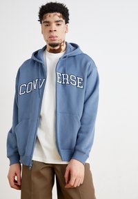 Felpa zip-up blu realizzata in morbido cotone misto, con la scritta "CONVERSE" in lettere bianche sul petto, tasca a marsupio e cappuccio regolabile con coulisse.