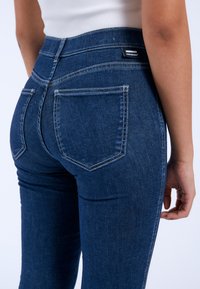 High-waisted blue denim jeans with four pockets, visible stitching, and a branded tag on the back waistband. Flared leg design.