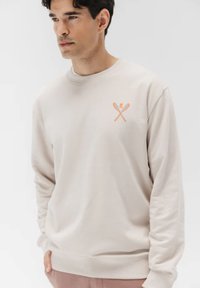 Sudadera de color beige claro con cuello redondo, mangas largas y un gráfico de esquí cruzado en naranja en el pecho. Material de algodón suave.