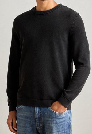 Mann trägt einen schwarzen, strukturierten Langarmpullover mit Rundhalsausschnitt und hat eine Hand in der hellblauen Jeans-Tasche vor einem schlichten Hintergrund.
