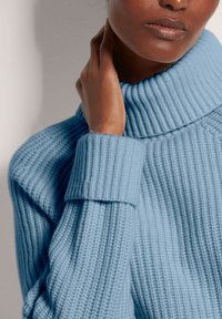 Person, der einen hellblauen, gerippten Rollkragenpullover trägt und sanft den Hals berührt.