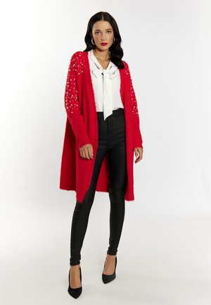 Cardigan rosso con decorazioni in perle sulle maniche, abbinato a una camicetta bianca e leggings in pelle nera. La modella indossa tacchi a punta neri.