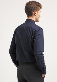 Donkerblauwe langemouwshirt met een gladde, glanzende afwerking, met een klassieke kraag en knoopmanchetten, gezien van achteren. Grijze pantalon eronder.