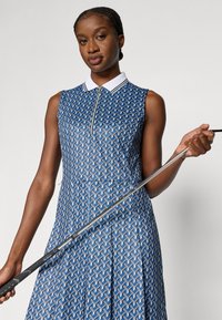 Zils bezroku kleita ar ģeometrisku rakstu, rāvējslēdzēja apkakli, baltām uzsvarām un krokotu svārku; modele tur golfa nūju.