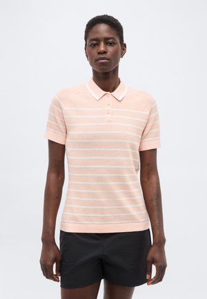 Naine kannab heledat roosat ja valget triibulist lühikeste varrukatega polosärki ning musti lühikeste pükse, seisab tavalise heleda tausta ees.