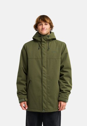 Jeune homme aux cheveux bruns courts portant une veste d'hiver verte à capuche et debout devant un fond blanc uni.