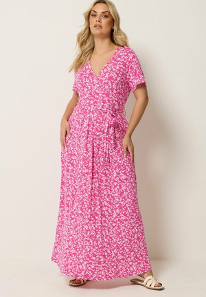 Kvinde står iført en lyserød omslagskjole med hvidt blomsterprint og korte ærmer, parret med hvide flade sandaler, mod en ensfarvet baggrund.