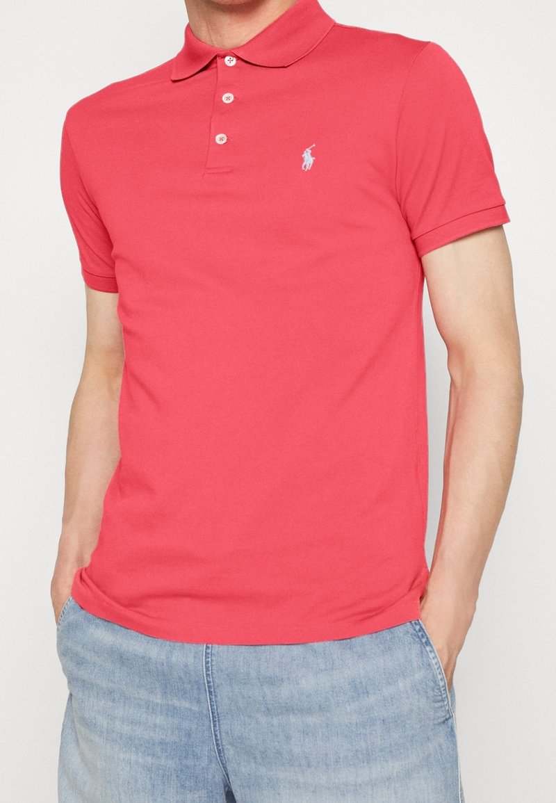 Homme portant un polo rouge à manches courtes avec trois boutons et un jean bleu clair, les mains dans les poches.