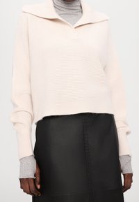 Hellbeiger gerippter Strickpullover mit weitem Kragen und kurzem Saum, kombiniert mit einem grauen Rollkragenpullover und einem schwarzen Lederrock.