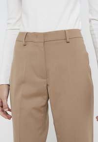 Pantalons marron clair en tissu texturé, avec un devant plat, des passants de ceinture et une coupe ajustée. Portés avec un haut à manches longues blanc.