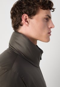 Doudoune marron foncé avec un col haut et une texture lisse et brillante. Le design présente des coutures piquées et une coupe ajustée.
