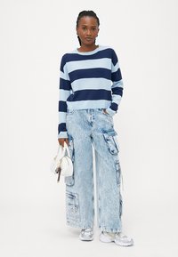 Suéter cropped de malha às riscas azul e marinho, combinado com calças de ganga cargo de perna larga em azul claro com bolsos, usado com ténis metálicos e uma mala branca.