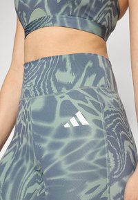 Un reggiseno sportivo e leggings a vita alta in un motivo astratto verde e grigio. Presenta il logo Adidas in bianco e un tessuto liscio e elasticizzato.