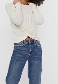 Maglione lavorato a maglia color crema con dettaglio annodato sul davanti, abbinato a jeans blu a vita alta. Il maglione presenta una texture a coste e maniche lunghe.