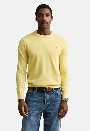 Mann trägt einen gelben Pullover mit langen Ärmeln, blaue Jeans mit schwarzem und braunem Gürtel sowie eine silberne Armbanduhr, steht vor weißem Hintergrund.